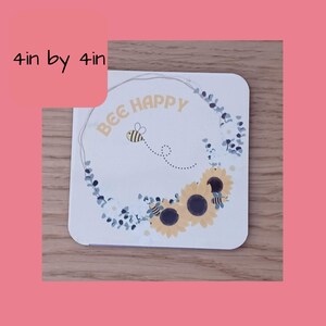 K&ouml;nnte beinhalten: Ein quadratischer Untersetzer in Wei&szlig; mit einem Kranz aus blauen Eukalyptusbl&auml;ttern und gelben Sonnenblumen. Eine Biene mit schwarzen und gelben Streifen fliegt im Kreis um den Text "BEE HAPPY".