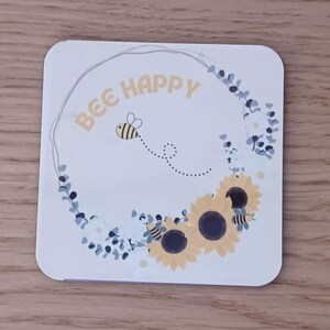 K&ouml;nnte beinhalten: Eine wei&szlig;e Karte mit einem Kranz aus blauen Eukalyptusbl&auml;ttern und wei&szlig;en Bl&uuml;ten. Der Kranz umgibt den Text "BEE HAPPY" in gelben Buchstaben. Eine gelbe und schwarze Biene fliegt &uuml;ber dem Text mit einer gepunkteten Linie, die hinter ihr herl&auml;uft. Drei gelbe Sonnenblumen mit schwarzen Zentren befinden sich am unteren Rand der Karte.