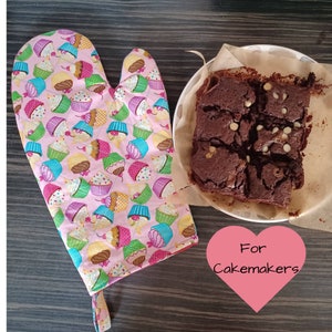 May include: Pink oven glove with a cupcake pattern beside a plate of brownies. The glove has a quilted design with colourful cupcakes on a pink background. The brownies are cut into squares and have white chocolate chips.