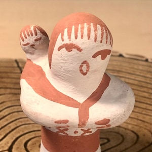 以下が含まれることがあります： 親が子供を抱いている様子を描いた陶器製の置物。人物は白とテラコッタ色で塗装されており、シンプルな顔の特徴と様式化された衣服が特徴です。ベースは明るい茶色です。