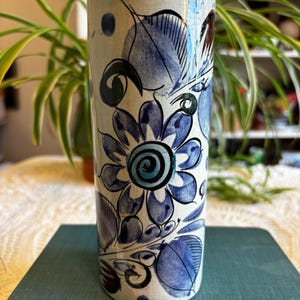 Puede incluir: Un jarrón cilíndrico de cerámica con base crema, adornado con patrones florales y de hojas azules. El jarrón presenta un gran diseño floral central y mide aproximadamente 20 cm de alto.