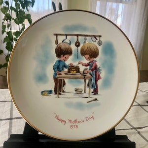 Op de afbeelding: Decoratieve plaat met een gouden rand met een illustratie van twee kinderen die een taart versieren. De plaat heeft de tekst "Happy Mother's Day 1978" in rode letters. De achtergrond is wit met een lichtblauwe lucht.