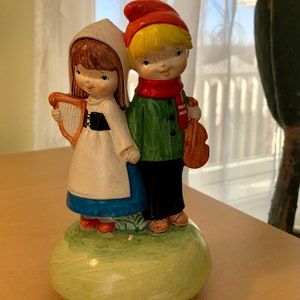 May include: A vintage ceramic figurine depicting a boy and girl in traditional attire. The girl holds a harp, and the boy carries a violin. They stand on a green, rounded base. The boy wears a red hat and scarf, and the girl wears a white bonnet.