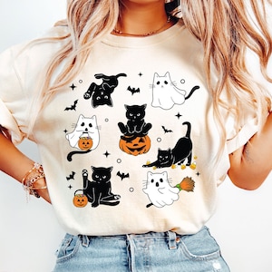 以下が含まれることがあります： ハロウィン用のコスチュームを着た猫の白黒のデザインが施された白いTシャツ。猫は、幽霊、魔女、カボチャなど、さまざまなコスチュームを着ています。デザインはシャツに複数回繰り返されています。