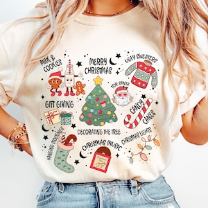Può includere: Una maglietta bianca con un design a tema natalizio che presenta illustrazioni di latte e biscotti, un albero di Natale, una calza, una canna di zucchero, un disco di Natale e Babbo Natale. Il testo sulla maglietta recita "Milk & Cookies", "Merry Christmas", "Ugly Sweaters", "For Santa", "Gift Giving", "Hanging Stockings", "Decorating the Tree", "Christmas Music", "Candy Canes" e "Christmas Lights".