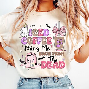 May include: A white t-shirt with a pink, purple, and brown graphic design featuring a ghost, a cup of iced coffee, a tombstone, and bats. The text on the shirt reads "Iced Coffee Bring Me Back From The Dead".