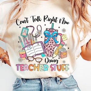 May include: A white t-shirt with the text "Can't Talk Right Now Doing Teacher Stuff" in colorful letters. The design includes a pink and black leopard print coffee cup, a stack of books, a pencil, and other school supplies.