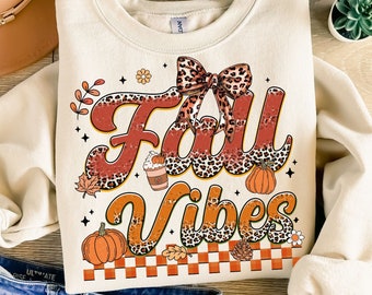 May include: A beige sweatshirt with a graphic print of the words "Fall Vibes" in a retro style. The graphic features a leopard print pattern, a bow, a pumpkin, and a cup of coffee.