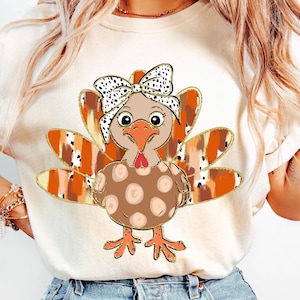 May include: A white t-shirt with a cartoon turkey wearing a brown and white polka dot bow. The turkey has a brown body with orange and brown feathers. The feathers have a gold glitter outline.