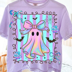 Puede incluir: Camiseta lila con un dise&ntilde;o de fantasma de dibujos animados, con un gran lazo, murci&eacute;lagos y flores. El fantasma est&aacute; enmarcado sobre un fondo a rayas. La camiseta est&aacute; colgada de una percha de madera.