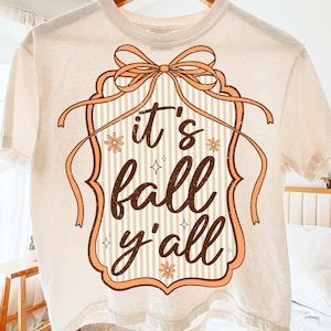 May include: Cream-colored t-shirt with the phrase "it's fall y'all" in a decorative frame. The frame is outlined in a peach color with a bow at the top and vertical stripes in the background. The text is in a brown script font.
