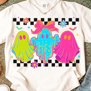 May include: Cream-coloured t-shirt with a vibrant graphic of three ghost characters. The ghosts are in green, blue, and pink, set against a black and white chequered pattern. The design includes flowers, stars, and bat motifs.