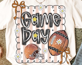 May include: Cream-colored t-shirt with a "Game Day" graphic. The design features a football, helmet, and daisies, set against a pink and white striped background. Brown bows and a black outline frame the design. A gold chain necklace is visible.
