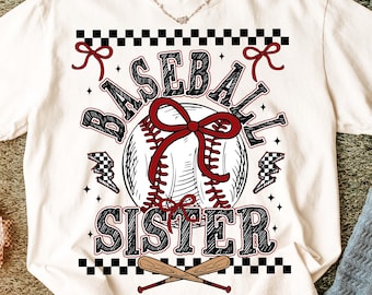 May include: Cream-colored t-shirt with a baseball-themed graphic. The design features a baseball with a red bow, the words "BASEBALL SISTER," and crossed baseball bats. Accents include black and white checkered patterns and lightning bolts.
