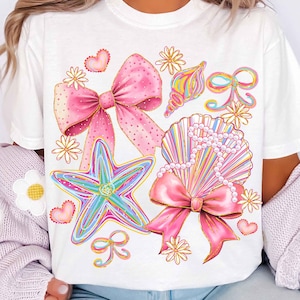 Op de afbeelding: Wit T-shirt met een kleurrijk ontwerp van zeeschelpen en zeesterren. Het ontwerp bevat roze strikken, harten en gouden accenten. Het shirt heeft een ronde hals en korte mouwen, ideaal voor casual wear.