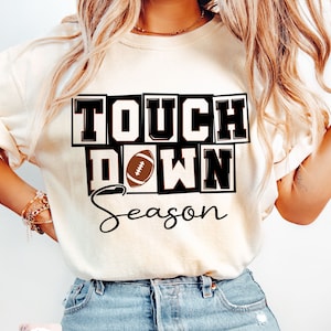 May include: A white t-shirt with the text "Touch Down Season" in black and brown block letters. A brown football is in the middle of the text.