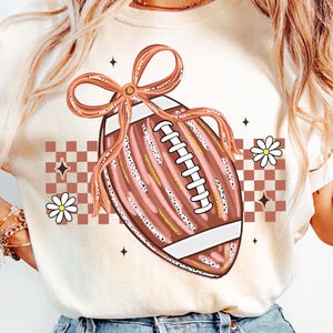 May include: Cream-colored t-shirt featuring a football design with a pink bow, white laces, and gold accents. The design includes a checkered pattern, daisies, and star accents, creating a sporty and feminine aesthetic.