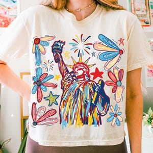 May include: A white t-shirt with a colorful graphic print of the Statue of Liberty, fireworks, and flowers. The graphic is in a cartoon style with bold lines and bright colors.