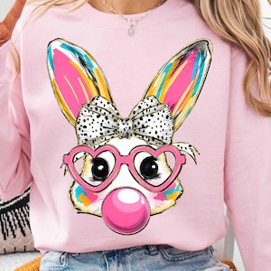 May include: Pink sweatshirt with a graphic of a cartoon bunny wearing heart-shaped sunglasses and a bow. The bunny is blowing a pink bubble.