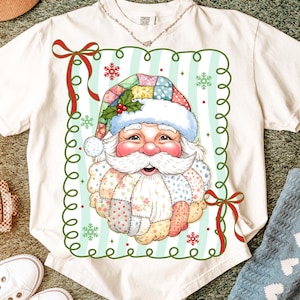 May include: Cream-colored t-shirt featuring a Santa Claus graphic. The Santa design has a patchwork hat and beard, with a smiling face. The graphic is framed with green swirls and red bows, perfect for the holiday season.