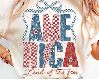 Op de afbeelding: Crèmekleurig T-shirt met een patriottisch ontwerp. Het woord "AMERICA" is weergegeven in rood, wit en blauw, met sterren en een dambordpatroon. De zin "Land of the free" staat eronder.