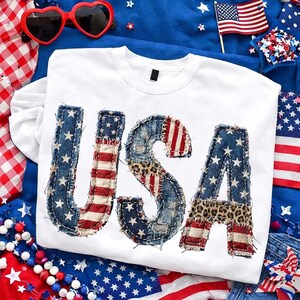 May include: White long-sleeved shirt with the word "USA" in a patchwork design. The letters feature denim, red and white stripes, leopard print, and blue fabric with white stars. Patriotic theme with heart-shaped sunglasses and an American flag.
