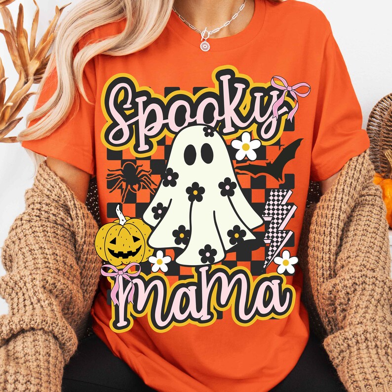 Puede incluir: Camiseta naranja con un dise&ntilde;o de Halloween. El dise&ntilde;o presenta un fantasma con flores, las palabras "Spooky Mama", una ara&ntilde;a, una calabaza y rayos. La camiseta tiene un estilo retro y vintage.