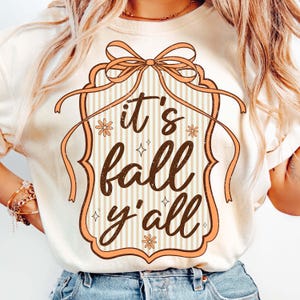May include: Cream-colored t-shirt with a fall-themed graphic. The design features the phrase "it's fall y'all" in a brown, decorative font, framed by a peach-colored border with a bow at the top. The background has vertical stripes.