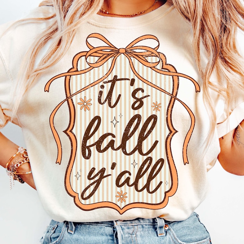 May include: Cream-colored t-shirt with a fall-themed graphic. The design features the phrase "it's fall y'all" in a brown, decorative font, framed by a peach-colored border with a bow at the top. The background has vertical stripes.