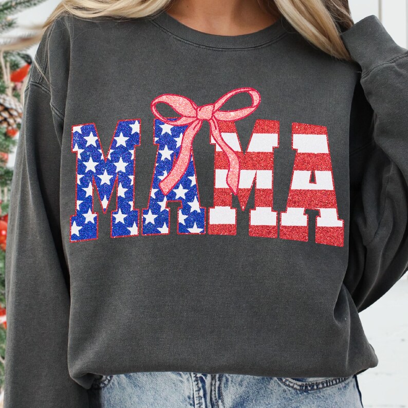May include: A charcoal gray sweatshirt with a red, white, and blue "MAMA" graphic. The letters are outlined in white and have a glitter effect. A pink bow is centered above the word.