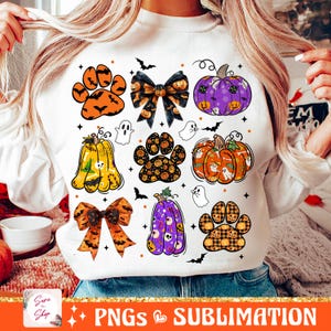 Puede incluir: Una sudadera blanca con un diseño de temática de Halloween con huellas de patas, calabazas, lazos y fantasmas. El diseño es en naranja, morado, negro y amarillo.