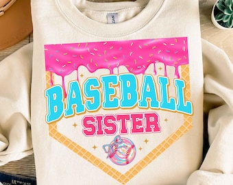 May include: Cream-colored sweatshirt with a graphic design. The design features the words "BASEBALL SISTER" in blue and pink, set within a baseball diamond shape. A pink donut with sprinkles tops the design, and a baseball with a bow is at the bottom.