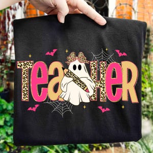 May include: Black t-shirt with a Halloween-themed design. The word "Teacher" is displayed in pink and tan leopard print lettering. A ghost wearing a leopard print bow and holding a pencil is in the center, with bats and spiderwebs.