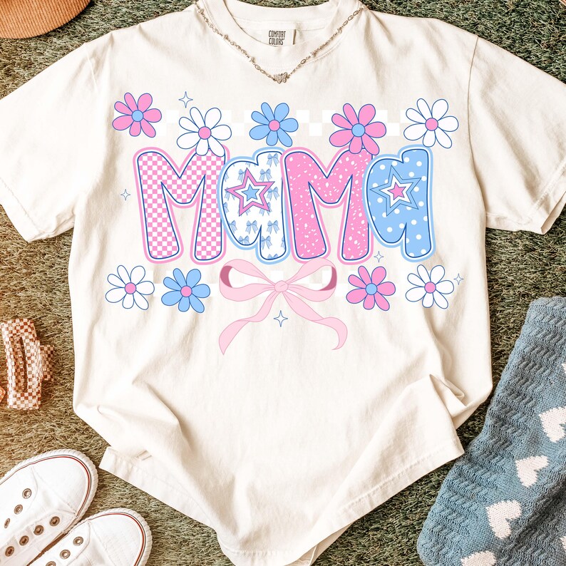 May include: Cream-colored t-shirt with the word "Mama" in a playful, colorful design. The letters are decorated with pink and blue patterns, stars, and flowers. A pink bow is below the word, and the shirt has a relaxed fit.