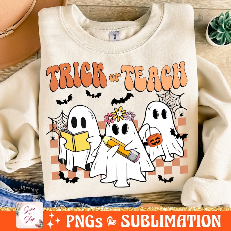 May include: A beige sweatshirt with the text "Trick or Teach" in orange and pink letters. Three ghosts are depicted, one holding a book, one holding a pencil, and one holding a pumpkin.