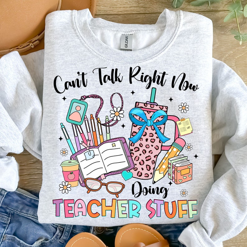 May include: A gray sweatshirt with the text "Can't Talk Right Now Doing Teacher Stuff" in rainbow colors. The text is surrounded by illustrations of school supplies, including a coffee cup, a book, a pencil, and a pair of glasses.