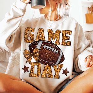Puede incluir: Sudadera color crema con las palabras "GAME DAY" en letras doradas con purpurina. Un bal&oacute;n de f&uacute;tbol americano marr&oacute;n con detalles plateados y un lazo dorado est&aacute; centrado en la camiseta. Tambi&eacute;n se ven dos estrellas doradas con purpurina.