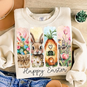 May include: A beige sweatshirt with a watercolor design featuring a bunny, a house, and flowers. The text "Happy Easter" is printed below the design.