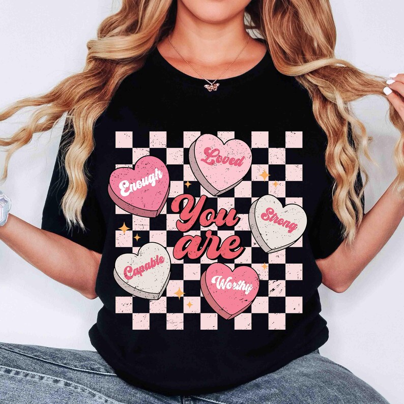 May include: Black t-shirt with a checkered pattern and pink hearts that say "Loved", "Enough", "Strong", "Capable", and "Worthy". The text "You are" is written in a retro font.