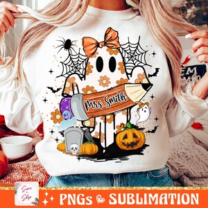 Puede incluir: Una sudadera blanca con un fantasma sosteniendo un l&aacute;piz que dice "Mrs. Smith". El fantasma est&aacute; rodeado de decoraciones de Halloween naranjas y negras, incluyendo calabazas, murci&eacute;lagos y telara&ntilde;as.