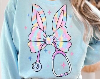 May include: A light blue sweatshirt with a tie-dye bunny ear bow and a stethoscope. The bow is pink, blue, and yellow with a rainbow tie-dye pattern. The stethoscope is pink and has a blue earpiece.