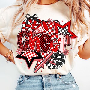 May include: A cream-colored t-shirt featuring a vibrant "Cheer" graphic. The design incorporates red, black, and white elements, including stars, pom-poms, and a megaphone. The word "Cheer" is stylized with various patterns, including leopard print and checkered designs.