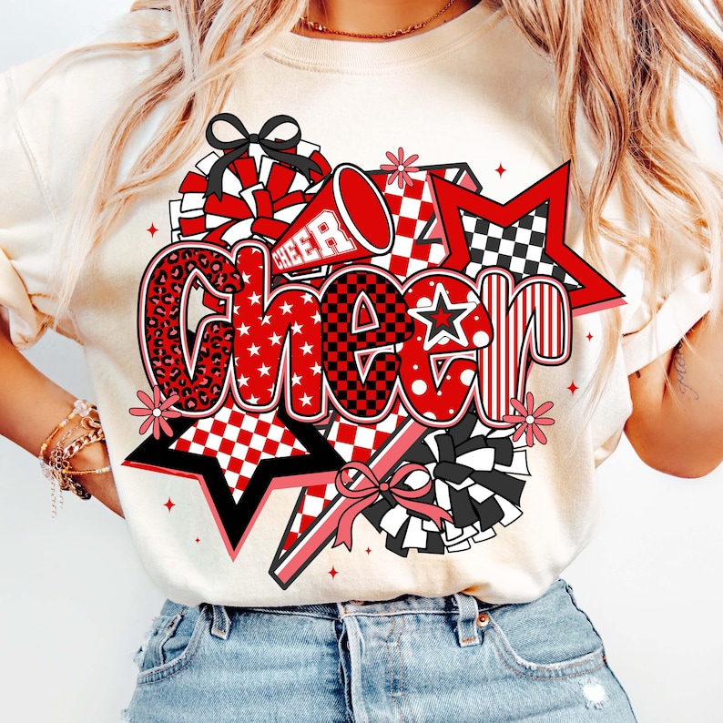 May include: A cream-colored t-shirt featuring a vibrant "Cheer" graphic. The design incorporates red, black, and white elements, including stars, pom-poms, and a megaphone. The word "Cheer" is stylized with various patterns, including leopard print and checkered designs.