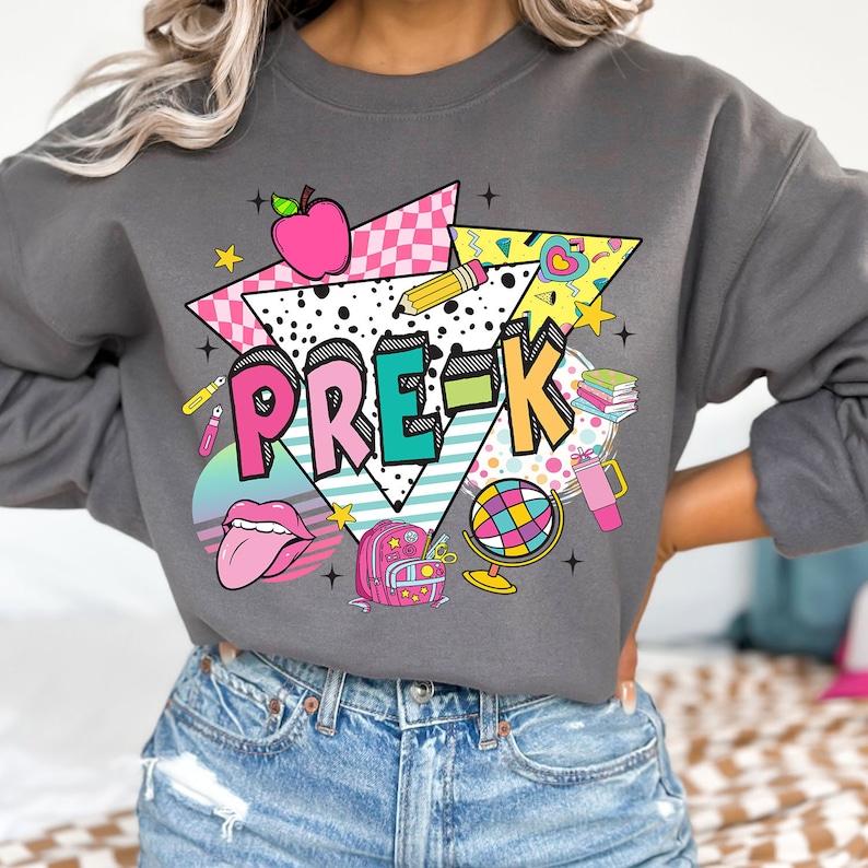 K&ouml;nnte beinhalten: Ein graues Sweatshirt mit einem bunten Grafikdesign, das den Text "PRE-K" zeigt, umgeben von Schulutensilien, einem Globus und einem Rucksack. Das Design ist im Retro-Stil der 80er Jahre.