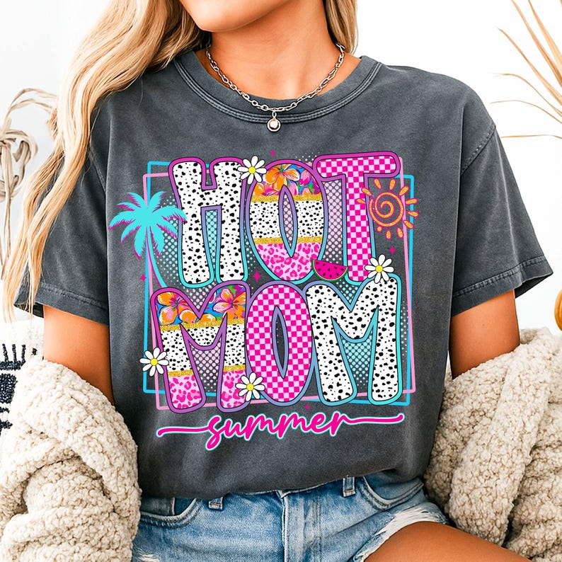 Puede incluir: Camiseta gris oscuro con un gr&aacute;fico colorido que dice "HOT MOM summer". El dise&ntilde;o incluye una palmera, flores y un sol. Las letras est&aacute;n rellenas de patrones como estampado de leopardo y dise&ntilde;os a cuadros.