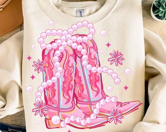 May include: Cream-colored sweatshirt featuring a pink graphic of cowboy boots adorned with a pearl necklace, flowers, and stars. The boots have light blue accents and are set against a cream background. A casual, country-themed design.
