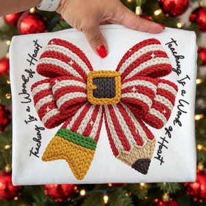 May include: White rectangular pillow with a knitted bow and pencil design. The bow is red and white striped with a gold buckle. The pencil has a yellow body and a brown tip. Text reads "Teaching Is a Work of Heart."
