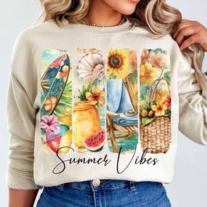 May include: A beige sweatshirt with a graphic print of summer-themed illustrations. The print features a surfboard, a seashell, a sunflower, a beach chair, a basket of fruit, and the text "Summer Vibes".