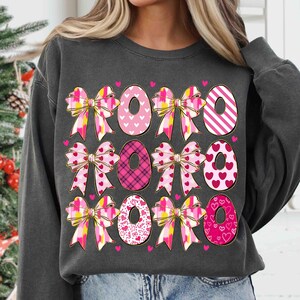 K&ouml;nnte beinhalten: Dunkelgraues Sweatshirt mit einem sich wiederholenden Muster aus Schleifen und den Buchstaben "XOXO". Die Schleifen sind rosa, gelb und wei&szlig;, w&auml;hrend die "O"s mit Herzen und Mustern verziert sind. Ein Design zum Valentinstag.