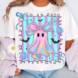 Puede incluir: Camiseta blanca con un dise&ntilde;o de fantasma de arco&iacute;ris pastel. El fantasma tiene un lazo y est&aacute; sobre un fondo a rayas. El dise&ntilde;o incluye murci&eacute;lagos, flores y una telara&ntilde;a, creando un tema de Halloween.